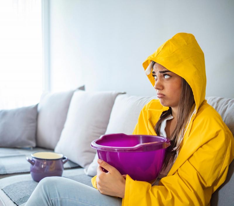 Eine Frau sitzt in einer nassen Wohnung auf einem Sofa in Magdeburg und hält eine Schüssel zum Auffangen von Tropfwasser.